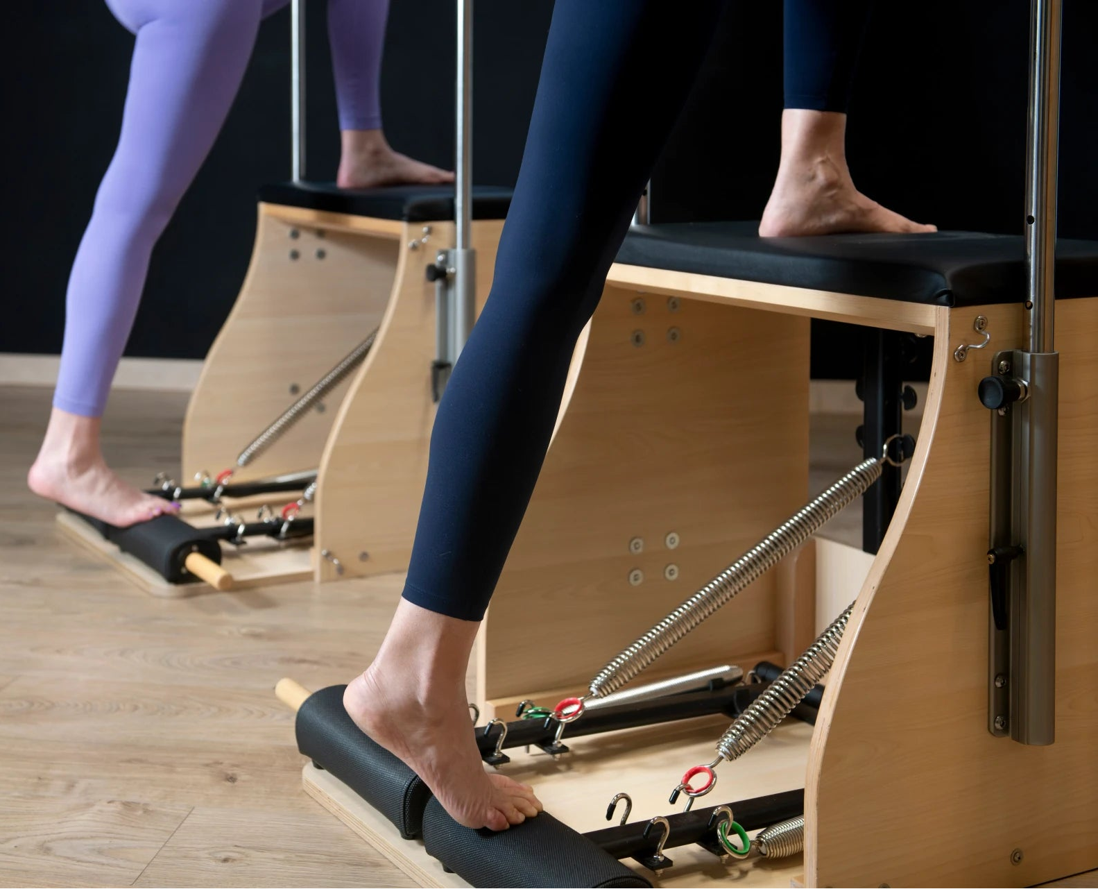 Pilates reformer machine in a studio with a person in navy leggings stepping onto the roller, another person in lavender leggings visible in the background, and a light wood floor beneath.