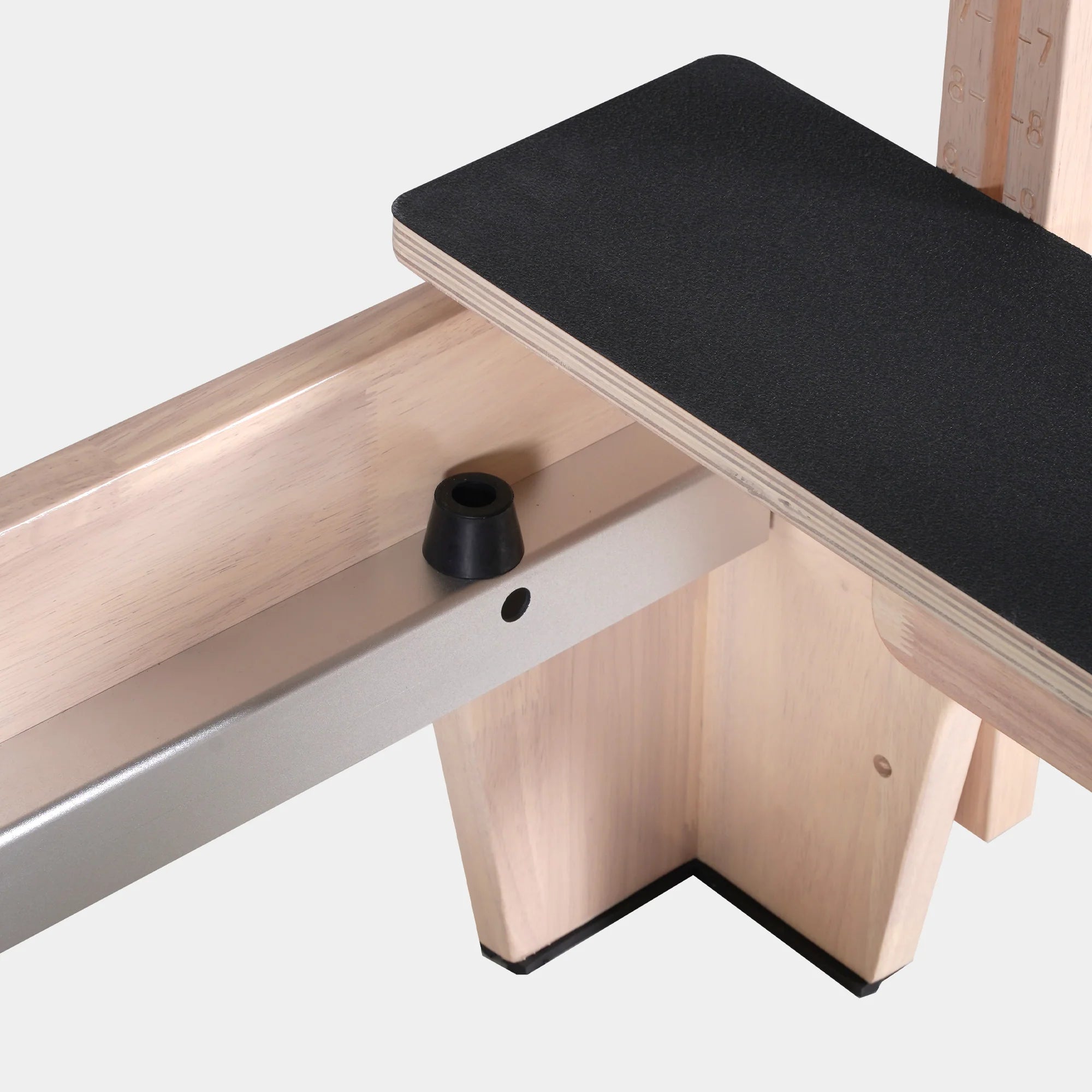 Close-up of a wooden exercise or massage table with a black textured top surface, light natural wood frame, and a silver metal support arm against a light gray background.